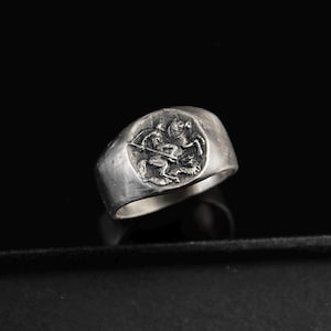 Può includere: Anello d'argento con un rilievo dettagliato di un cavaliere a cavallo, con una lancia. L'anello ha una faccia rettangolare arrotondata e una fascia spessa. L'anello è esposto su uno sfondo scuro.