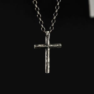 Puede incluir: Collar con colgante de cruz plateada. La cruz tiene un aspecto texturizado y rústico. El fondo es negro, lo que resalta la joya.