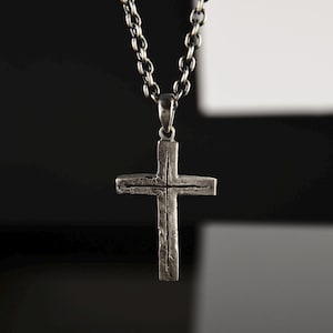 Puede incluir: Un collar de cadena de plata con un colgante de cruz de plata. La cruz tiene una superficie texturizada y está ligeramente elevada.