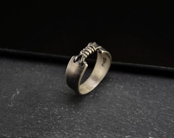 May include: A silver ring with a textured band and a twisted wire design. The ring is on a dark grey surface.