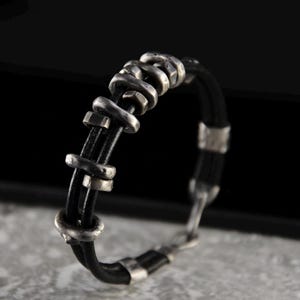 以下が含まれることがあります： 銀色の金属製のアクセントが付いた黒い革のブレスレット。ブレスレットは編み込まれた革のストラップと複数の銀色の金属部品を持っています。
