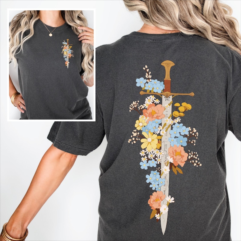 A woman wearing a charcoal grey Comfort Colors t-shirt. On the front left chest (pocket area), there is a small, vertical illustration of a vintage sword intertwined with colorful wildflowers like daisies and forget-me-nots. The back of the shirt