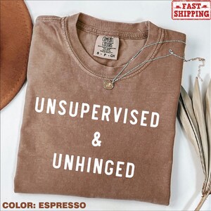 May include: Espresso brown t-shirt with the text "UNSUPERVISED & UNHINGED" in white.