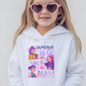 May include: White hooded sweatshirt featuring a colorful graphic with the text "HUNTRAX RUMI MIRA ZOLY". The graphic includes illustrations of three cartoon characters. The sweatshirt has a front pocket and a drawstring hood.
