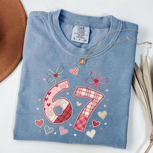 Puede incluir: Camiseta azul claro Comfort Colors con los números 67 aplicados en un diseño de patchwork. Los números están decorados con corazones y lazos. Un collar dorado con un colgante está sobre la camiseta.