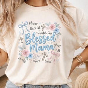 Puede incluir: Camiseta color crema con las palabras "Blessed Mama" en escritura azul, rodeada de gráficos florales y cruces. Palabras adicionales incluyen "Mama", "Grateful", "Thankful", "Joy", "Family", "Peace", "Loved", "Heart" y "Full".