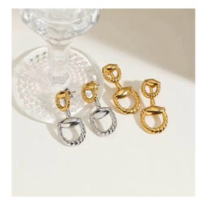 Puede incluir: Pendientes colgantes dorados y plateados. Presentan un diseño único con una parte superior circular y una parte inferior circular más grande y texturizada. Los pendientes se muestran sobre una superficie blanca, con un objeto de vidrio transparente en el fondo.