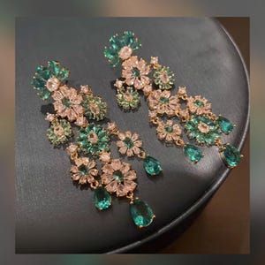 Peut inclure: Boucles d'oreilles dorées élégantes avec un motif floral orné de cristaux verts et transparents. Les boucles d'oreilles ont un design en cascade avec des pendentifs en cristal vert en forme de goutte. Sur fond sombre.