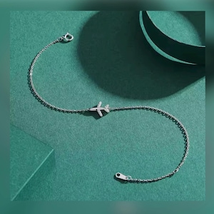 Peut inclure: Un bracelet avion argenté. Le bracelet présente un petit charme d'avion avec des détails étincelants. La chaîne est fine et élégante, avec un fermoir sécurisé. Le bracelet est présenté sur une surface verte.