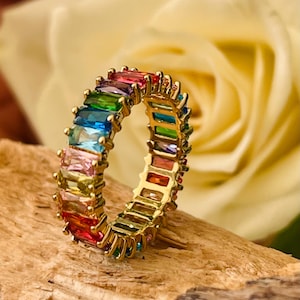 Op de afbeelding: Een goudkleurige ring met een band van kleurrijke, rechthoekige edelstenen. De edelstenen zijn gerangschikt in een regenboogpatroon, waaronder rood, oranje, geel, groen, blauw en paars. De ring ligt op een houten oppervlak.
