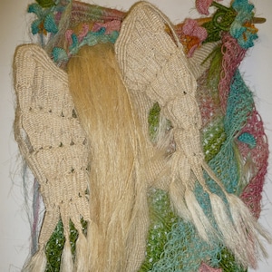 Può includere: Un arazzo da parete fatto a mano con una figura all'uncinetto con lunghi capelli biondi e ali. La figura è circondata da fiori e foglie all'uncinetto in tonalità di verde, blu, rosa e bianco.