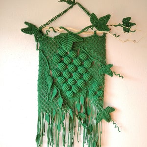 May include: Green macrame wall hanging featuring a grape design and leaves. The hanging is crafted from green cord and has a fringe at the bottom.