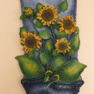 Könnte beinhalten: Ein Wandbehang mit einer gehäkelten Darstellung einer Topfpflanze mit vier Sonnenblumen. Die Sonnenblumen sind gelb mit braunen Zentren und die Blätter sind grün. Der Topf ist dunkelblau und der Hintergrund ist grau.