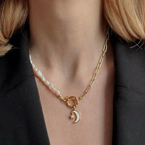 Op de afbeelding: Een goudkleurige ketting met een parel- en kettingontwerp. De ketting heeft een witte emaille maanvormige hanger met een steraccent. De ketting is vastgezet met een gouden sluiting.