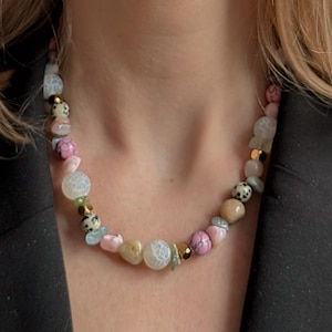 Könnte beinhalten: Eine farbenfrohe Perlenkette mit Edelsteinen in Rosa, Weiß, Beige und Grün. Die Halskette enthält runde und unregelmäßig geformte Perlen mit goldfarbenen Akzenten. Die Halskette wird über einer schwarzen Jacke getragen.
