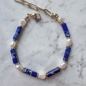 May include: A gold chain bracelet with blue and white beads. The blue beads are rectangular and have a marbled pattern. The white beads are round and resemble pearls.