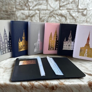 Puede incluir: Una colección de portapasaportes en varios colores, incluyendo blanco, azul marino, gris, rosa y negro. Cada uno presenta una imagen dorada o plateada de un templo. Un portapasaportes negro abierto con una tarjeta y papel en primer plano.