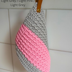 May include: A handmade, crocheted kitchen towel in light grey and light pink. The towel has a diagonal stripe of pink and a leather loop for hanging. The text "Light Grey / Light Pink / Light Grey" is visible.