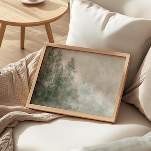 A cozy Japandi interior setting featuring a framed artwork of a misty forest on a textured beige and green background. The wooden frame rests gently against a soft beige couch, draped with a knit blanket, enhancing the warm and organic aesthetic. In the background, a round wooden table adds to the natural ambiance, while the neutral color palette and minimalist design create a harmonious, clutter-free space. Soft lighting and natural materials evoke a sense of calm and effortless elegance.
