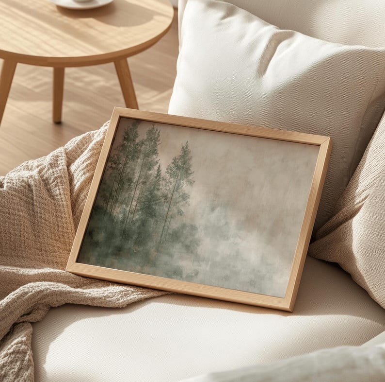 A cozy Japandi interior setting featuring a framed artwork of a misty forest on a textured beige and green background. The wooden frame rests gently against a soft beige couch, draped with a knit blanket, enhancing the warm and organic aesthetic. In the background, a round wooden table adds to the natural ambiance, while the neutral color palette and minimalist design create a harmonious, clutter-free space. Soft lighting and natural materials evoke a sense of calm and effortless elegance.