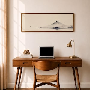 May include: A wooden desk with a laptop, books, and a gold desk lamp. A framed landscape painting of a mountain scene hangs above the desk. A wooden chair with a woven seat is in front of the desk. The room has a neutral colour palette.