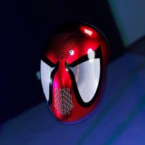 May include: A red and black Spider-Man mask with white mesh eye coverings. The mask has a glossy finish and is set against a dark blue background. The mask's design includes a textured pattern on the mouth area.