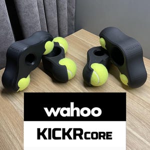 Op de afbeelding: Zwarte kunststof fiets trainer stand met vier gele tennisballen bevestigd. De stand heeft de tekst "Wahoo KICKR CORE" erop gedrukt.