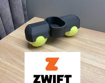 May include: A black plastic device with two tennis balls attached to the sides and a small cup in the centre. The device is labelled "ZWIFT" in orange and black lettering.