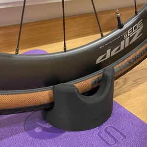 May include: A black bicycle wheel stand with a brown tyre and a black rim. The wheel is resting on a purple yoga mat. The wheel has a black valve stem and the text "DIZ SEIDE" printed on the side.