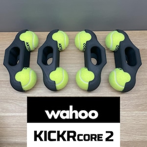 Könnte beinhalten: Vier schwarze Tennisballhalter mit gelben Tennisbällen. Die Halter haben ein einzigartiges Design mit einem Griff und einer gebogenen Form zur Sicherung der Bälle. Der Text "wahoo KICKR CORE 2" ist ebenfalls zu sehen.