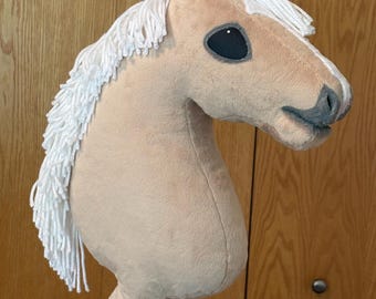 May include: A plush hobby horse with a tan fabric head, a white yarn mane, and a white yarn forelock. The horse features dark grey felt eyes and a grey stitched nose detail. The head is mounted on a wooden stick, creating a toy horse design.