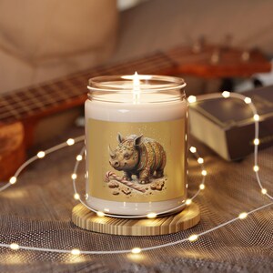 May include: A white candle with a gold label featuring a gingerbread rhinoceros with candy cane and sprinkles. The candle is on a wooden base and surrounded by string lights.