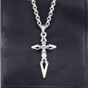 Peut inclure: Un collier pendentif argenté avec un design en forme de croix. La croix présente des extrémités pointues et un motif géométrique détaillé. La chaîne est à maillons et le fond est de couleur sombre.