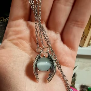 May include: A silver necklace with a pendant. The pendant has a light blue, spherical stone held by stylized wings. The chain is made of small links. The pendant is approximately 2.5 cm in diameter.