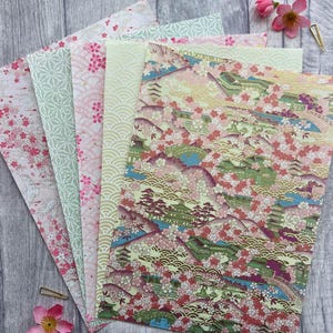 Könnte beinhalten: Ein Set aus sechs Dekorpapieren mit japanisch inspirierten Designs. Die Papiere sind in Grün-, Rosa- und Goldtönen gehalten und zeigen Muster von Kirschblüten, Wellen und geometrischen Formen.