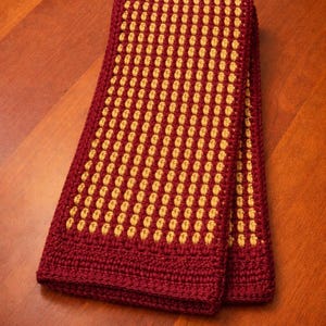Puede incluir: Una toalla rectangular doblada, tejida a mano. La toalla presenta un patrón repetitivo de puntos amarillo dorado sobre un fondo burdeos oscuro. El borde es de color burdeos liso. La toalla está sobre una superficie de madera.