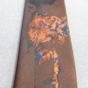 Puede incluir: Corbata marrón con un patrón abstracto en negro, naranja y azul. La corbata está hecha de una tela lisa y tejida. El diseño presenta formas y texturas irregulares. Fondo gris claro. Accesorio formal.