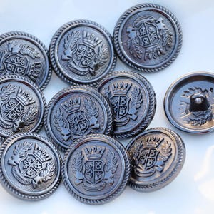 May include: Eight silver-toned buttons with a raised design of a crest with a crown and leaves. The buttons have a textured, rope-like border.