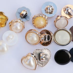 May include: Assortment of decorative buttons in various shapes, colors, and materials, including pearl, gold, silver, and black. Some buttons have a heart shape, others are round, and some are star-shaped.