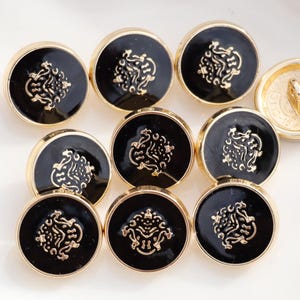 May include: A collection of black and gold buttons with an ornate design. The buttons have a glossy black center with a gold-colored decorative pattern and a gold-colored rim. One button is turned over, showing the back.