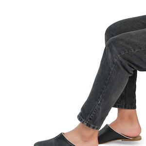 May include: Black leather slip-on shoes with a gold trim around the edge. The shoes are worn by a person wearing dark denim jeans.