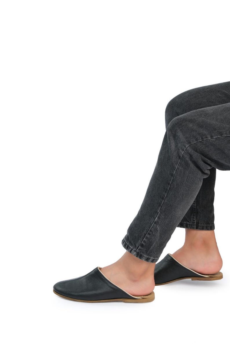 May include: Black leather slip-on shoes with a gold trim around the edge. The shoes are worn by a person wearing dark denim jeans.