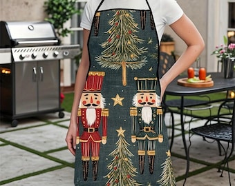 May include: A dark teal apron featuring a Christmas-themed design. The apron showcases two nutcracker figures in red and green uniforms, along with Christmas trees and gold stars. The apron has a black neck strap and is worn by a person.
