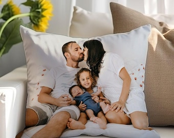 Könnte beinhalten: Quadratisches weißes Kissen mit einem Familienporträt. Das Bild zeigt eine vierköpfige Familie, wobei sich die Eltern küssen und ihre beiden Kinder. Das Kissen liegt auf einem weißen Sofa mit anderen Kissen im Hintergrund.