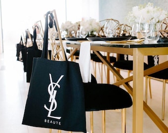 May include: Black tote bag with white "YSL BEAUTE" logo hanging on a gold chair. The image shows a table setting with black plates, gold cutlery, and white floral arrangements. The room has a modern, elegant aesthetic.