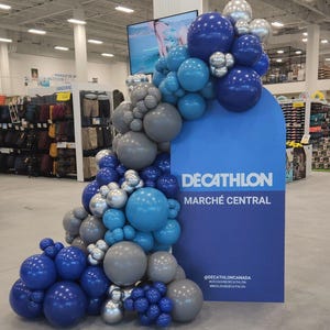 Puede incluir: Una exhibición en una tienda Decathlon presenta un letrero azul con el nombre de la tienda y un arco de globos. El arco está compuesto por globos azules, plateados y grises de varios tamaños, creando una disposición decorativa y llamativa.