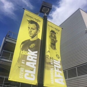 Puede incluir: Dos pancartas amarillas con retratos de jugadores cuelgan de una farola. Las pancartas presentan el texto "COLUMBUS MAPFRE STADIUM" y los nombres "CLARK" y "AFFUL" con sus números de camiseta. El cielo es azul con nubes.
