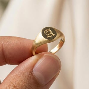 May include: A gold signet ring with a dark circular face featuring a stylized letter "A". The ring is held between fingers, showcasing its polished, metallic finish. The ring's design is simple and elegant, suitable for everyday wear.
