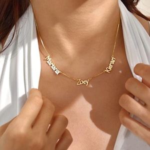 Op de afbeelding: Een goudkleurige ketting met drie naamhangers: "Mariana", "Zoey" en "Kieran". De namen zijn in een sierlijke lettertype. De ketting wordt gedragen om de nek van een persoon, tegen een witte top.