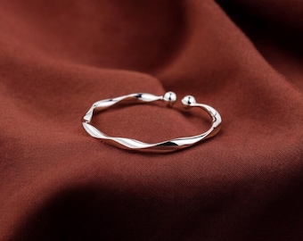 May include: A silver-coloured, open cuff bracelet with a twisted design. The bracelet has a smooth, reflective surface and is set against a dark red-brown background. The ends of the bracelet have small, round ball accents.